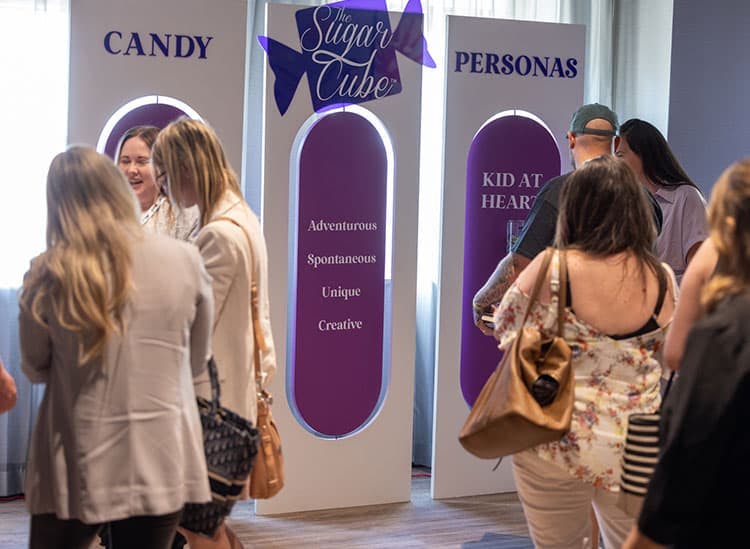  The image shows an indoor event space where attendees are interacting near two promotional standing banners. The banners are purple with white text; one reads 