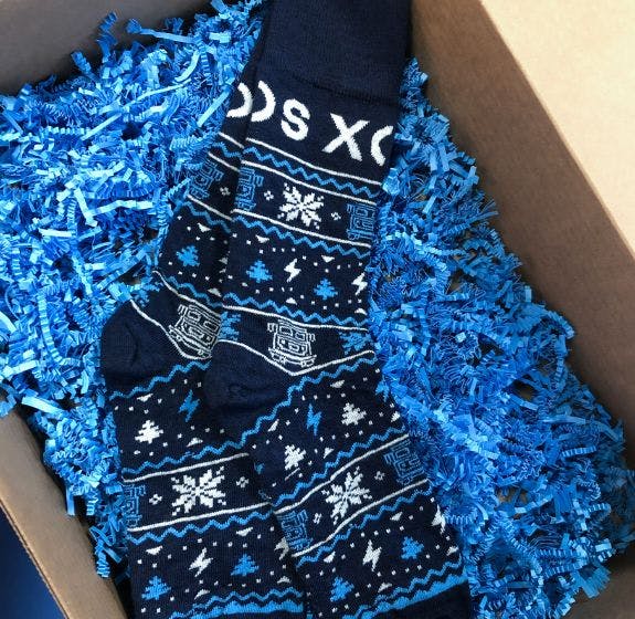 Custom branded holiday socks for Xos Trucks from Sock Club, featuring a blue winter themed design and blue crinkle paper in a box.