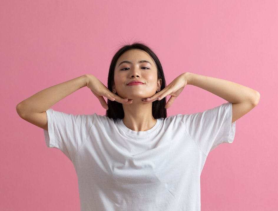 Millennial woman posing with her hands under her chin in a white t-shirt in front of a pink background