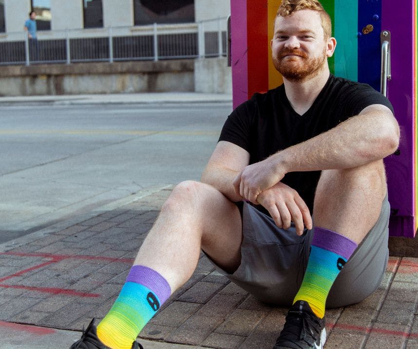 A main with red hair sitting on the street by a rainbow-painted pole with rainbow custom socks on his feet