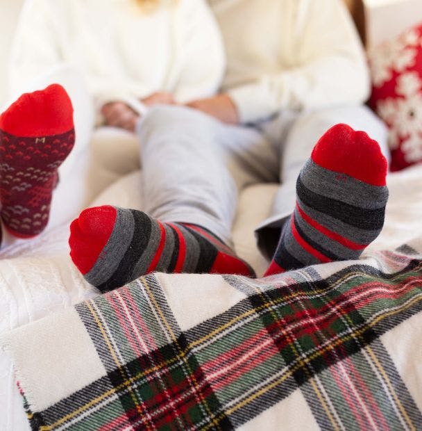 Custom striped wool socks being worn by a man lounging in a cozy bed with a plaid blanket and a lot of pillows