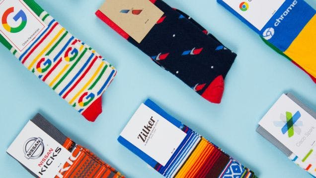 A lot of colorful custom socks with logos for brands including Google, American Airlines, Nissan, and Cisco on a light blue background