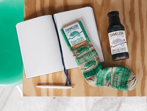 Green themed Chameleon Cold Brew Coffee socks paired with a coffee drink, notebook, and pen on a desk