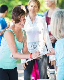 A woman at a 5k event for nonprofit fundraising signing up to give a donation on a clipboard with a sunny park in the background