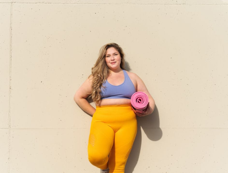 Plus size model in athletic yoga gear posing outside against a cream colored wall