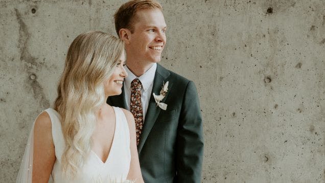 Bride Brittany and Groom Jonathan posing in their wedding outfits in front of a concrete wall