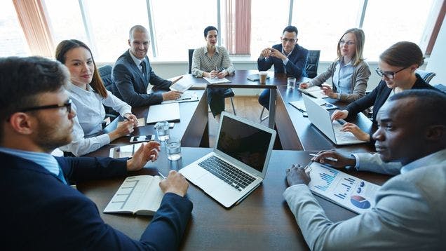 A corporate team sitting around a conference table at a corporate event with computers and charts in a modern conference room