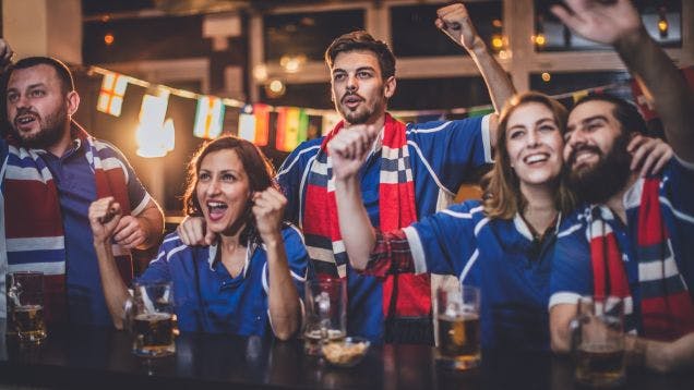 A group of soccer fans in sports merch cheering on their team at a bar with beers in front of them