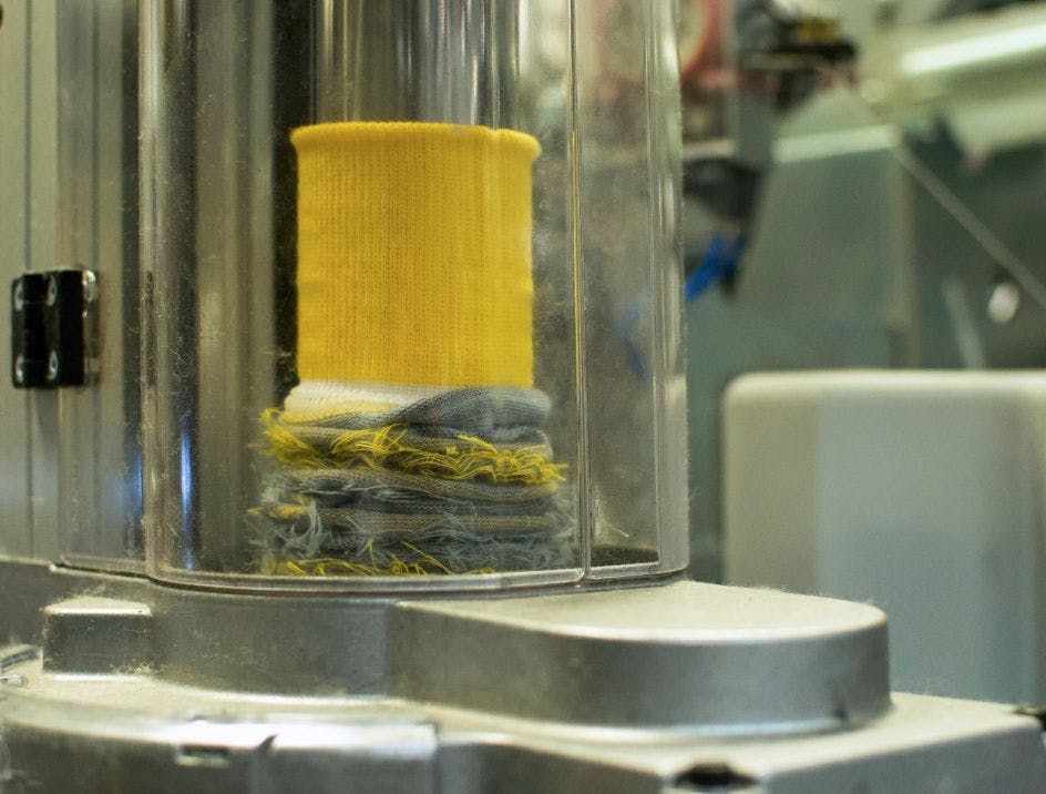 A yellow sock cuff being knit on a circular knitting machine.
