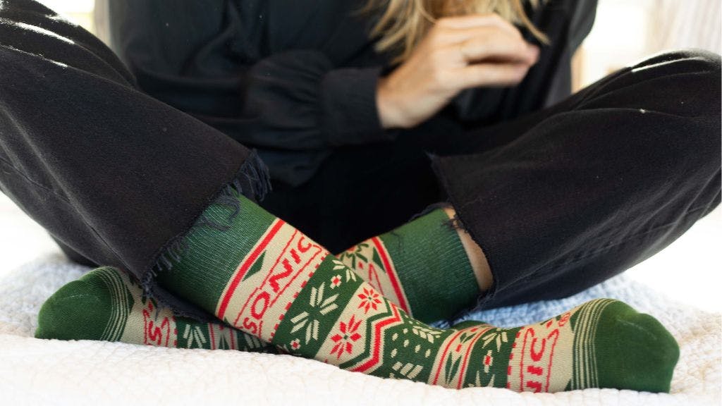 Woman sitting on a white bed wearing red and green branded socks for Sonic with a red and green Christmas sock design