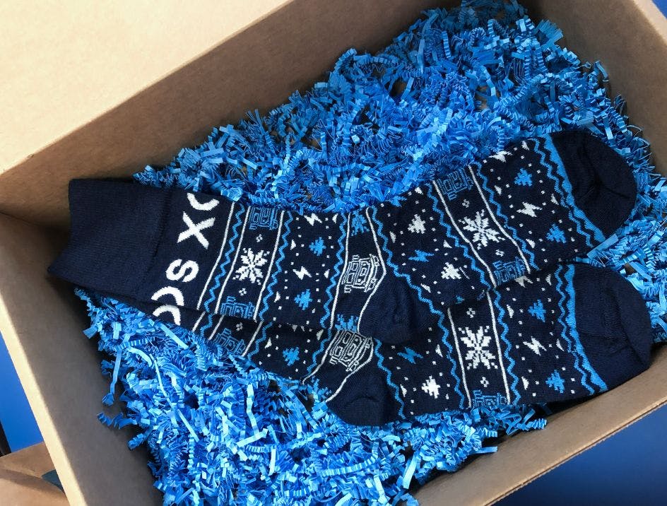 Custom branded holiday socks for Xos Trucks from Sock Club, featuring a blue winter themed design and blue crinkle paper in a box.