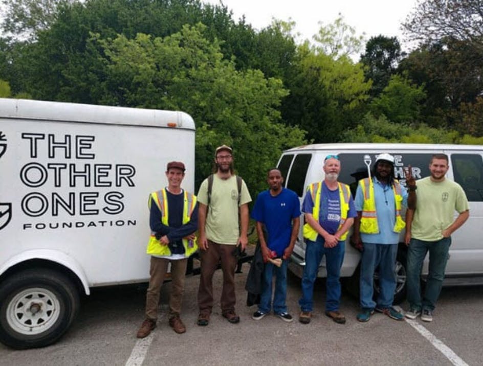 Group of men smiling wearing construction vests in front of white van and trailer.