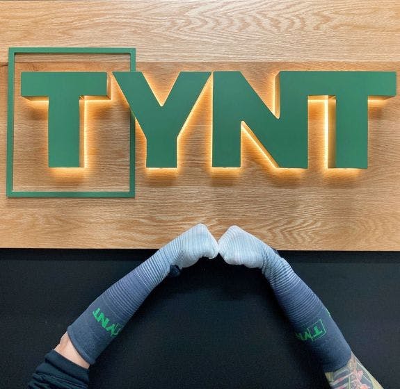 Two arms wearing Tynt custom socks in front of a wooden sign featuring the Tynt logo