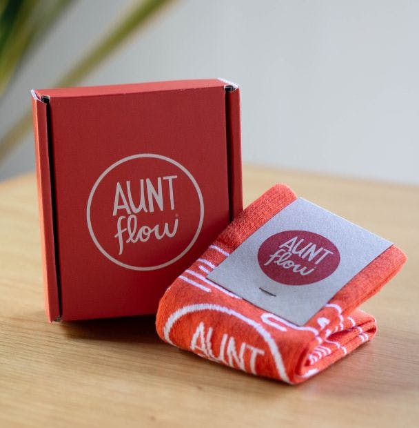 Custom mailer box for Aunt Flow sitting with a matching pair of branded Aunt Flow socks on a desk with a plant in the background