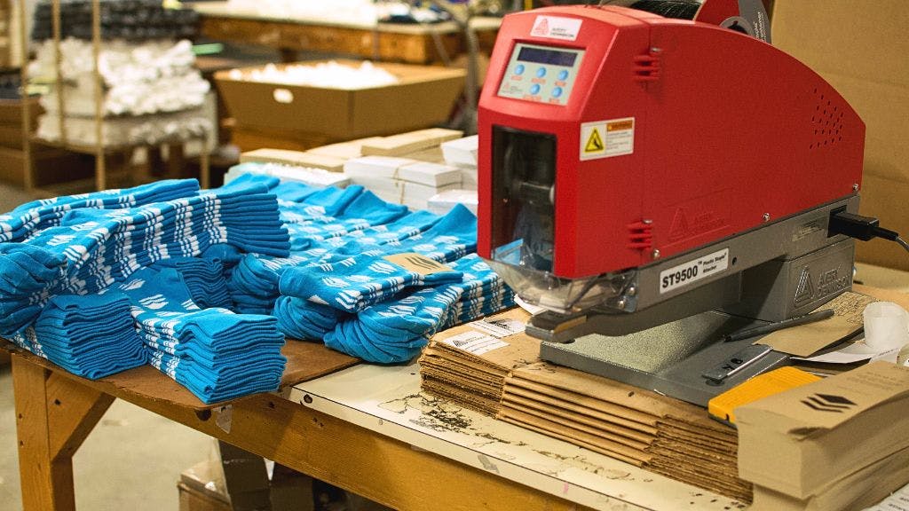A custom sock finishing and tacking machine sitting next to a pile of blue custom socks ready to be packaged