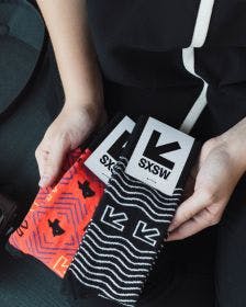 Hands holding custom socks for SXSW with custom header cards on a black background with a camera in the background