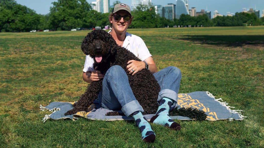 Man holding black dog sitting in grass wearing blue face socks