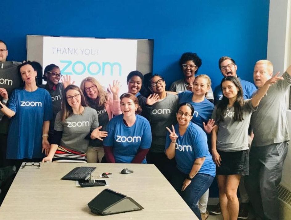 A group of Zoom employees in Zoom t-shirts in a blue conference room waving at the camera