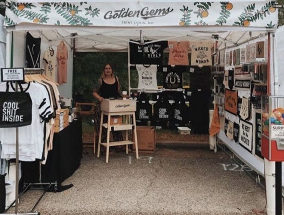 Golden Gems custom merch tent at an event showing t-shirts, banners, bandanas, and a person working the register.