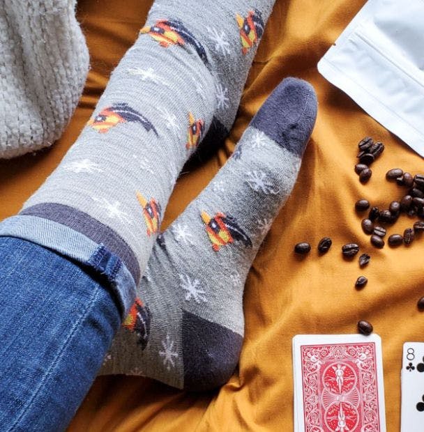 Custom grey wool socks with logo bring worn by a woman lounging in a cozy bed with coffee and playing cards