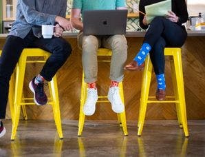 Three employees sitting on yellow stools in an office kitchen wearing branded socks