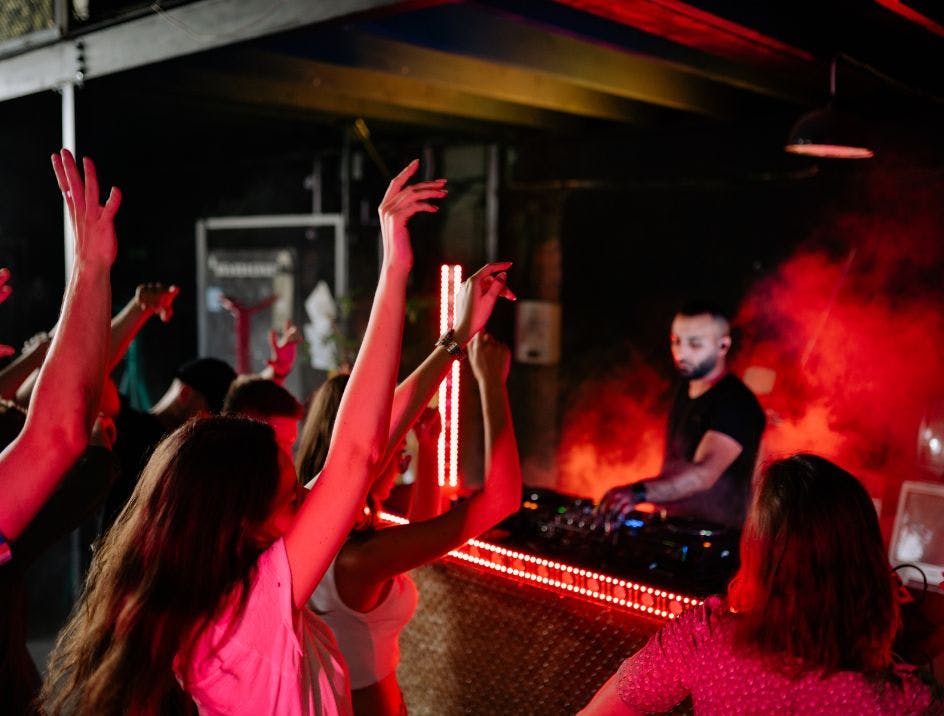 A DJ in a DJ booth with lights and fog playing in front of people dancing with their arms in the air at a company holiday party.