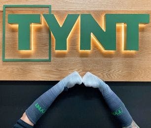 Two arms wearing Tynt custom socks in front of a wooden sign featuring the Tynt logo