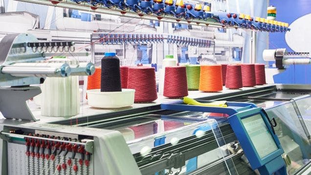 Knitting machine in a brightly lit knitting facility with colorful spools of yarn on top of the machine feeding into it