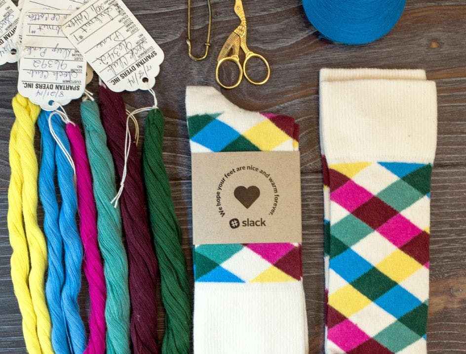 Custom socks for Slack on a wood table with yarn samples showing the yarn colors that were used in the custom sock design