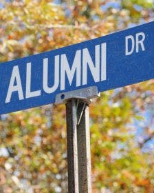 A blue street sign for Alumni Drive on a college campus with fall trees and leaves in the background and a blue sky
