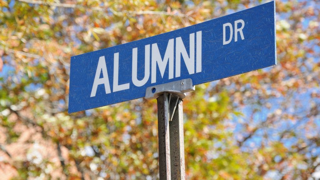 A blue street sign for Alumni Drive on a college campus with fall trees and leaves in the background and a blue sky