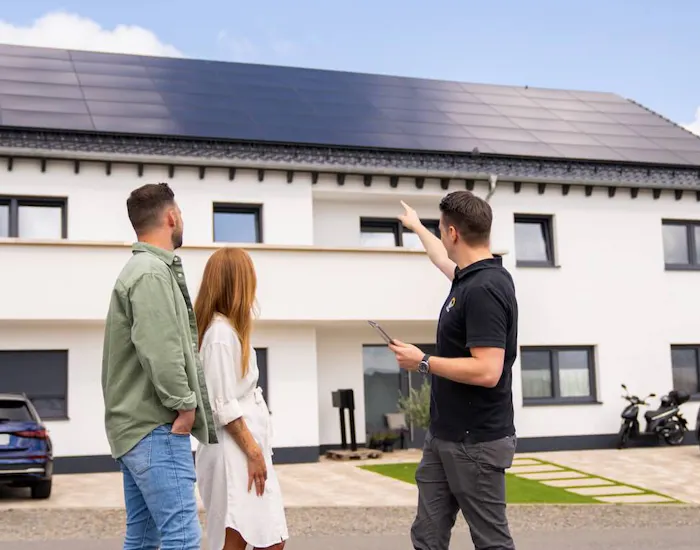 Paar und Photovoltaik Berater stehen vor Mehrfamilienhaus mit Solaranlage und unterhalten sich Paar und Photovoltaik Berater stehen vor Mehrfamilienhaus mit Solaranlage und unterhalten sich