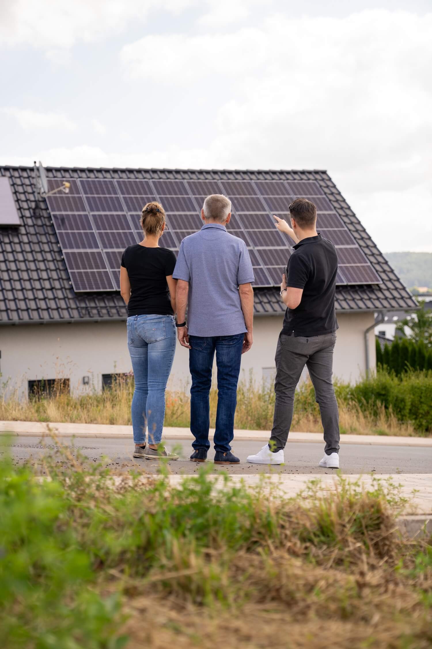 Paar und Solarberater stehen vor einem Wohnhaus und schauen auf die Solaranlage auf dem Dach