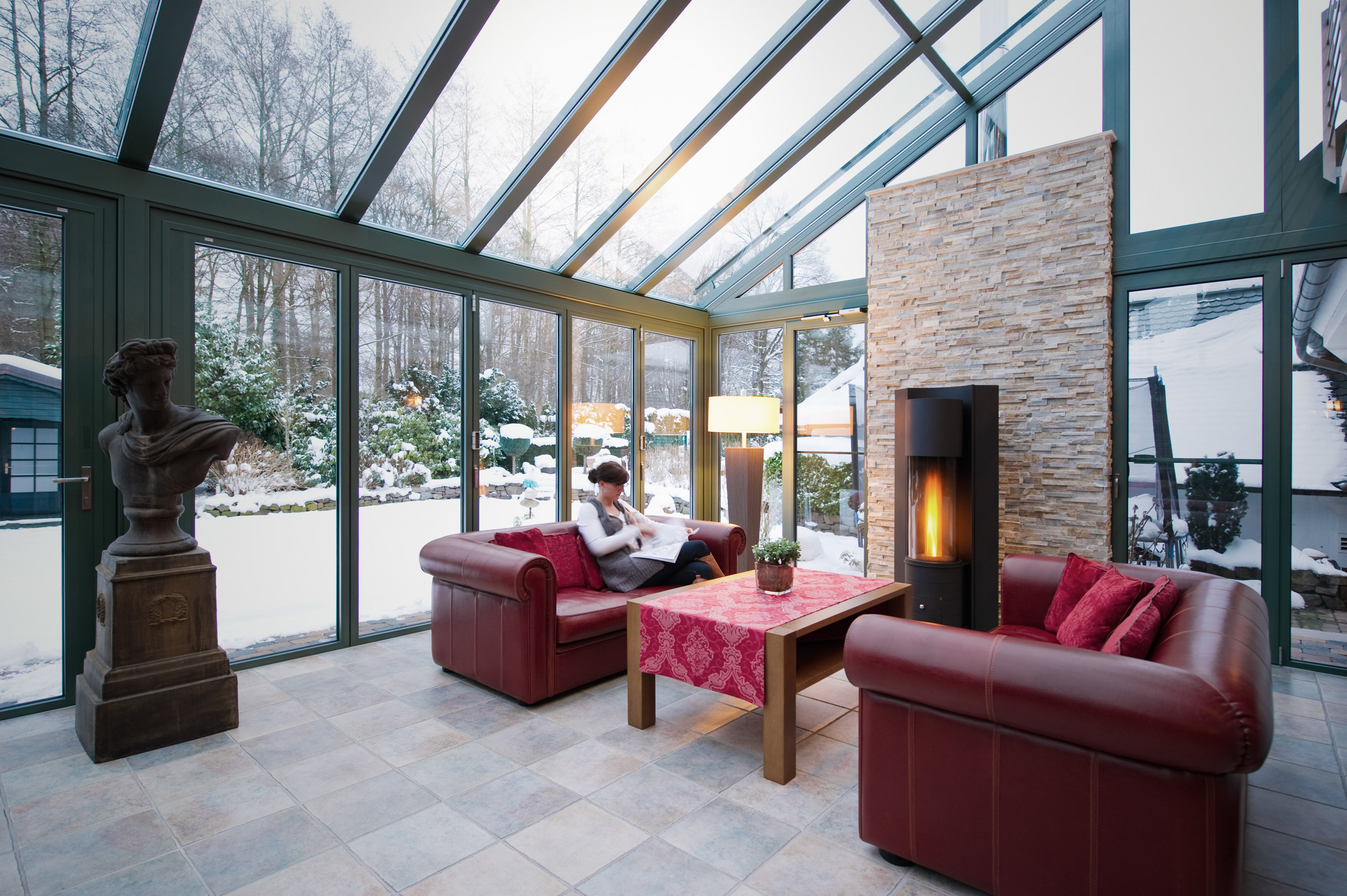 Lady reading on a red leather sofa next to log burner with snow outside 