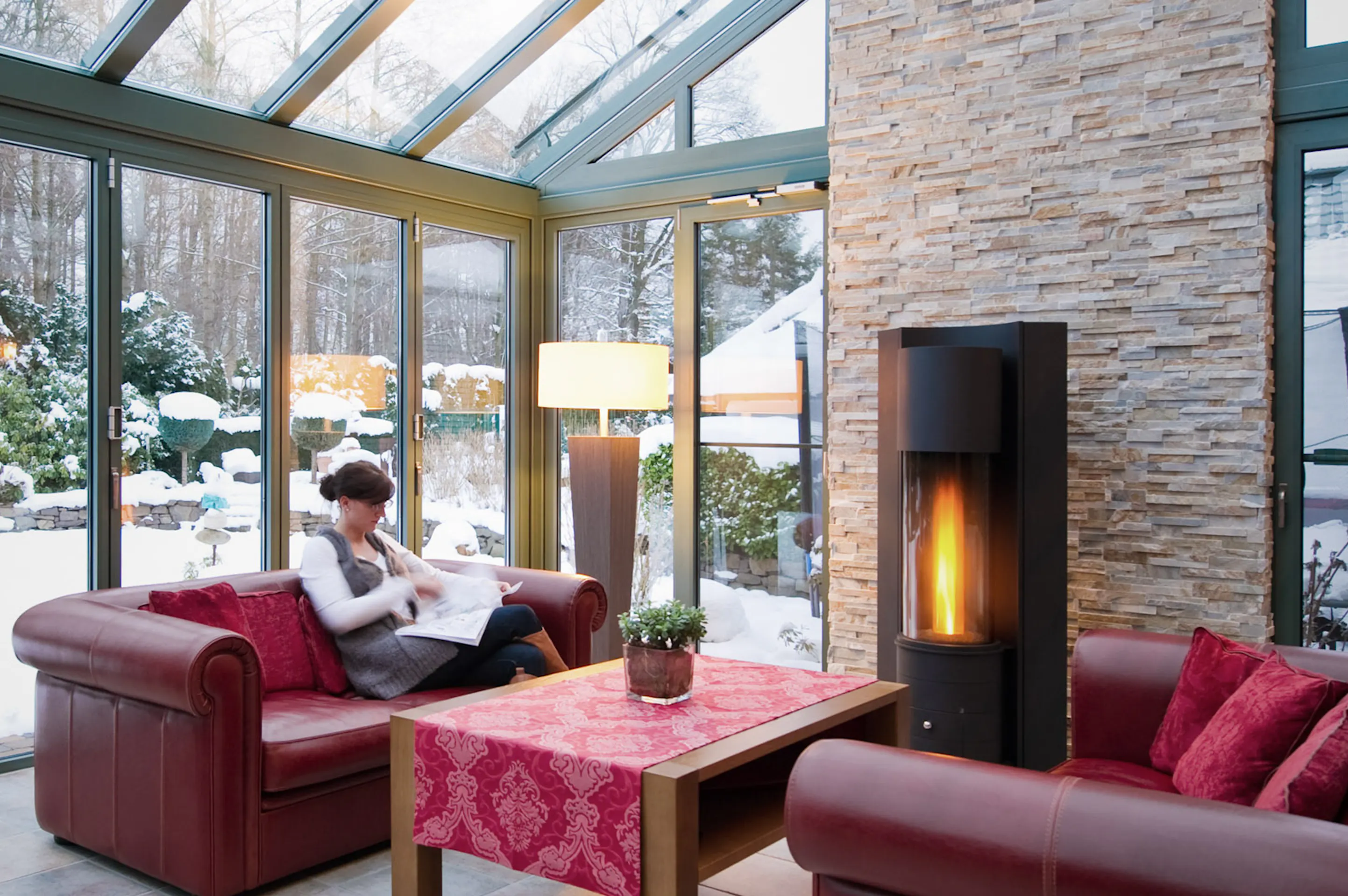 Lady reading on a red leather sofa next to log burner with snow outside