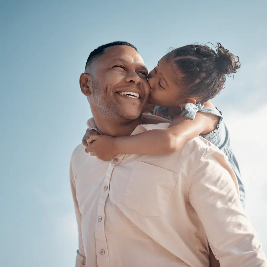 Pai sorrindo carregando filha nas costas enquanto ela beija o rosto dele
