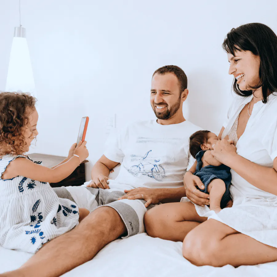 Família brincando e sorrindo deitados juntos na cama de casal com filha tirando foto do irmão ainda bebê amamentando no peito da mãe e pai sorrindo ao lado