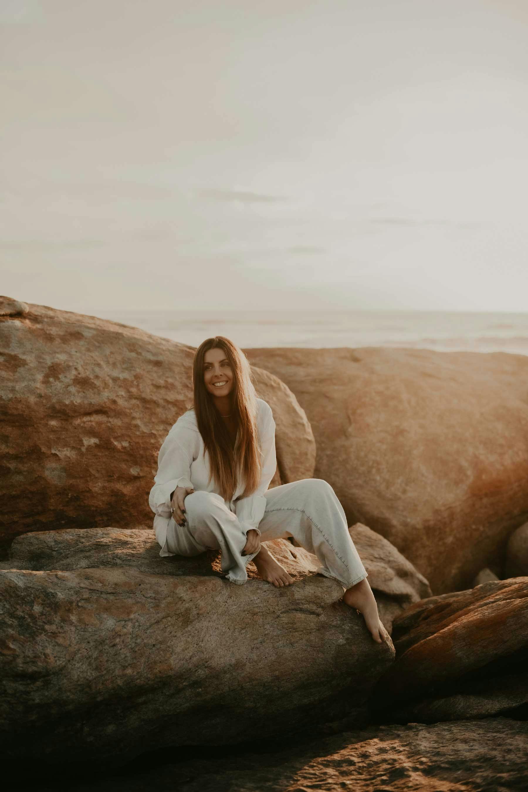 Our founder sitting on rocks by the ocean, smiling at the camera