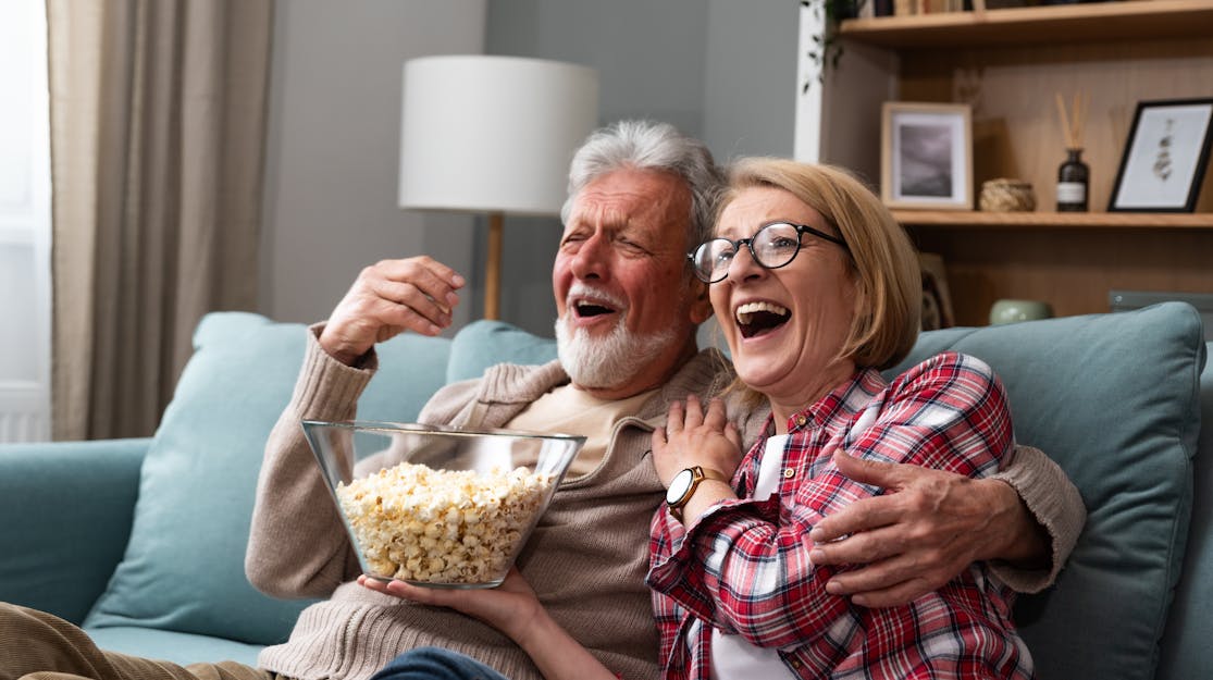 An elderly couple sitting on the lounge embracing sharing a bowl of popcorn, reacting to a film in front of them. 