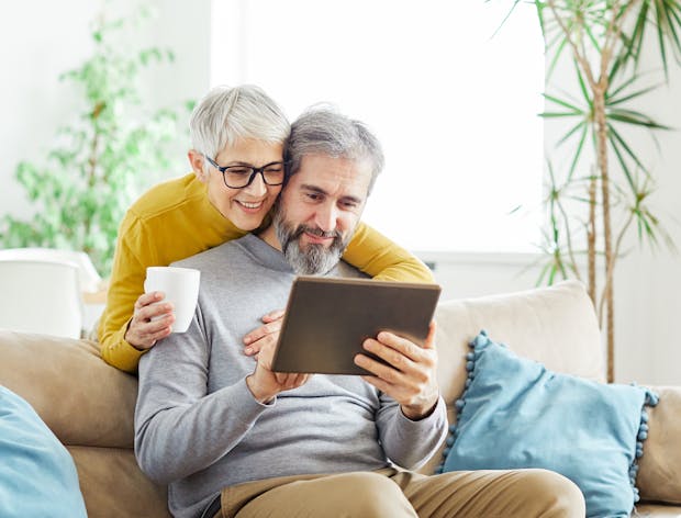 A man and woman looking at something amusing on a tablet device sitting on a beige coloured couch. 