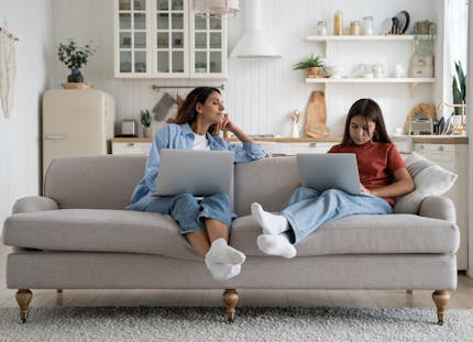 A mother and daughter sitting on the couch looking at their laptops. The mother is watching what her daughter is doing on her laptop. The couch is great and on a rug with a kitchen in the background. The image is using all neutral tones. 