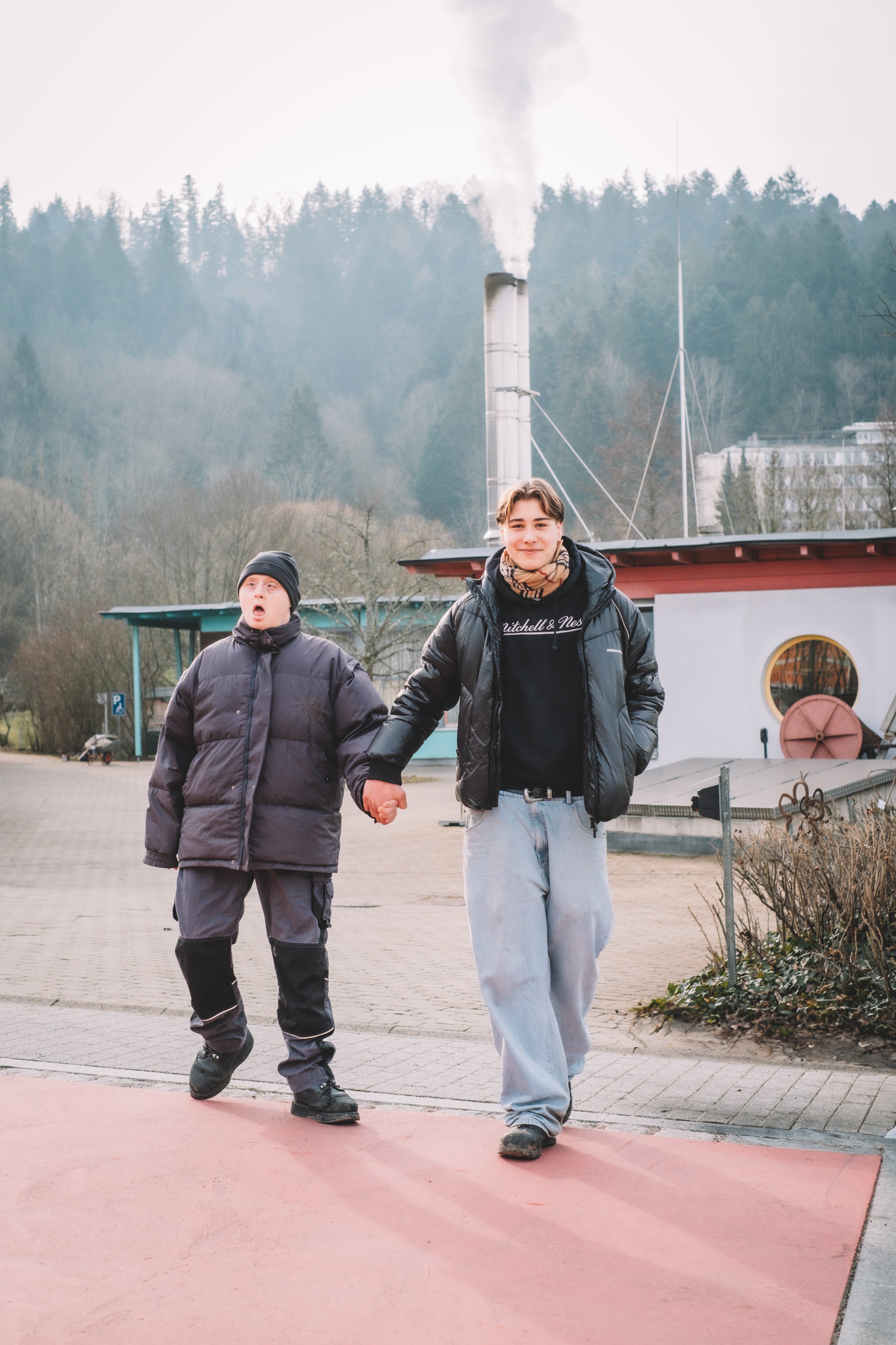 Man sieht zwei Männer, die sich an der Hand halten und auf einem roten Weg laufen. Der rechte Mann ist circa 16 Jahre alt und lächelt in die Kamera. Er geht circa einen Schritt vor dem anderen Mann. Dieser trägt eine Mütze und hat den Mund offen.