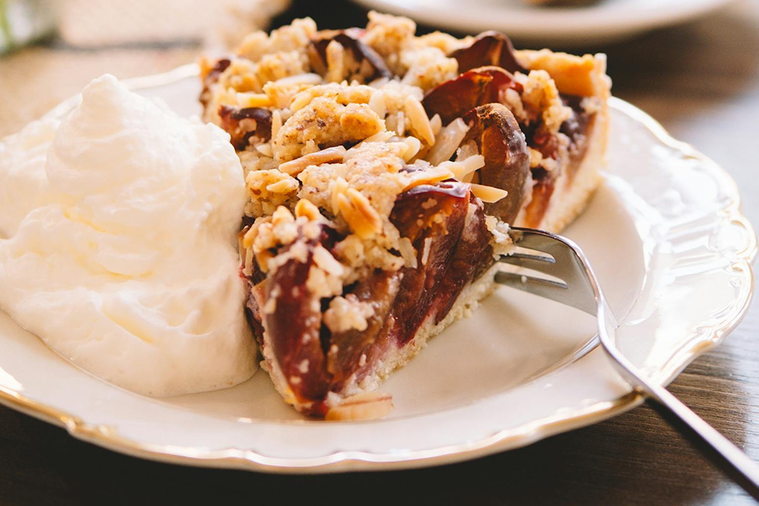 Man sieht einen Kuchen mit hellem Boden, Pflaumen und Streuseln. Der Kuchen steht auf einem weißen Teller. Es liegt eine silberne Gabel rechts neben dem Kuchen. Links neben dem Kuchen auf dem Teller liegt ein Kleks Schlagsahne.