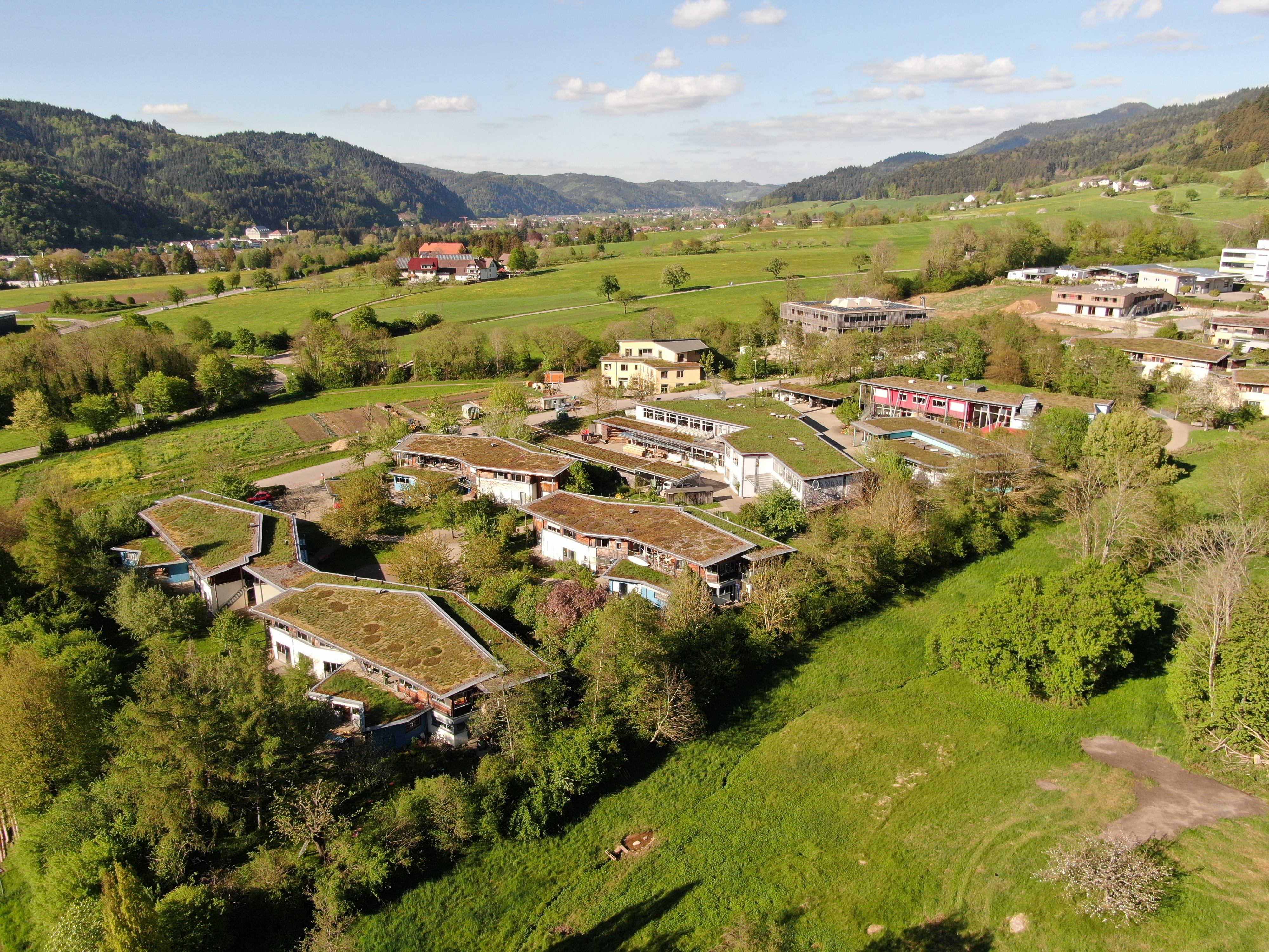 Es ist ein Drohnenbild mit mehreren Häusern in einem grünen Gelände zu sehen. Im Horizont sieht man blauen Himmel und Berge sowie Bäume.