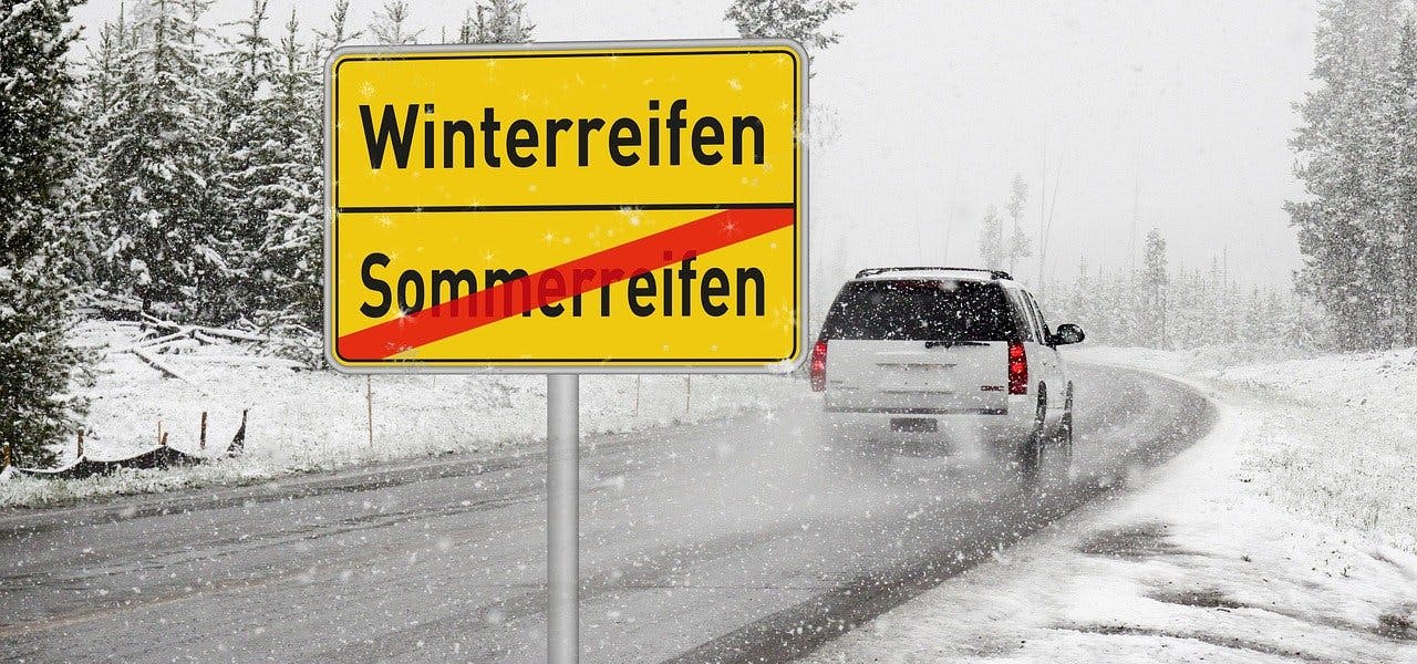 Sicher durch den Winter: Gutscheine für Winterreifen Auto fährt im Schneeregen an Schild vorbei, auf dem Sommerreifen durchgestrichen und Winterreifen angekündigt werden.