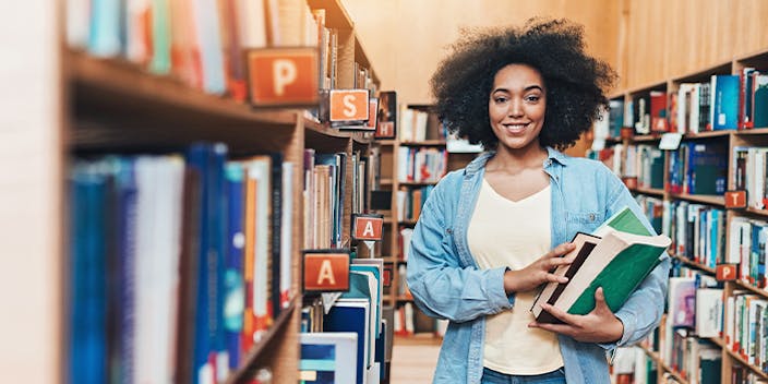 Starterkit für die Uni – Gutscheine für Schreibwaren, Möbel und Elektronik Schwarze Studentin hat Bücher in der Hand und steht zwischen Bücherregalen in der Bibliothek