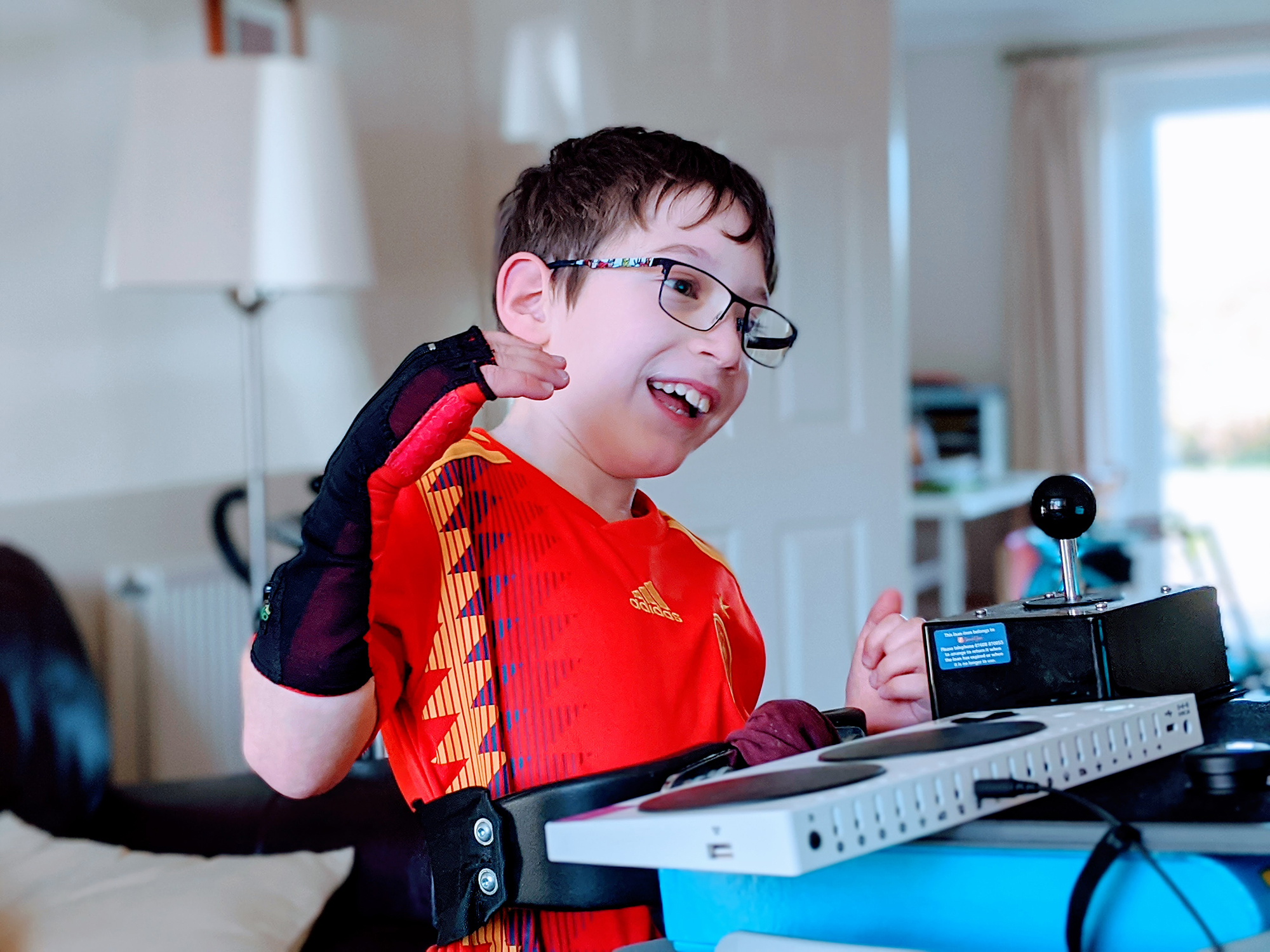 a young boy wearing a wrist brace and a red football tshirt plays with an xbox adaptive controller and a large joystick