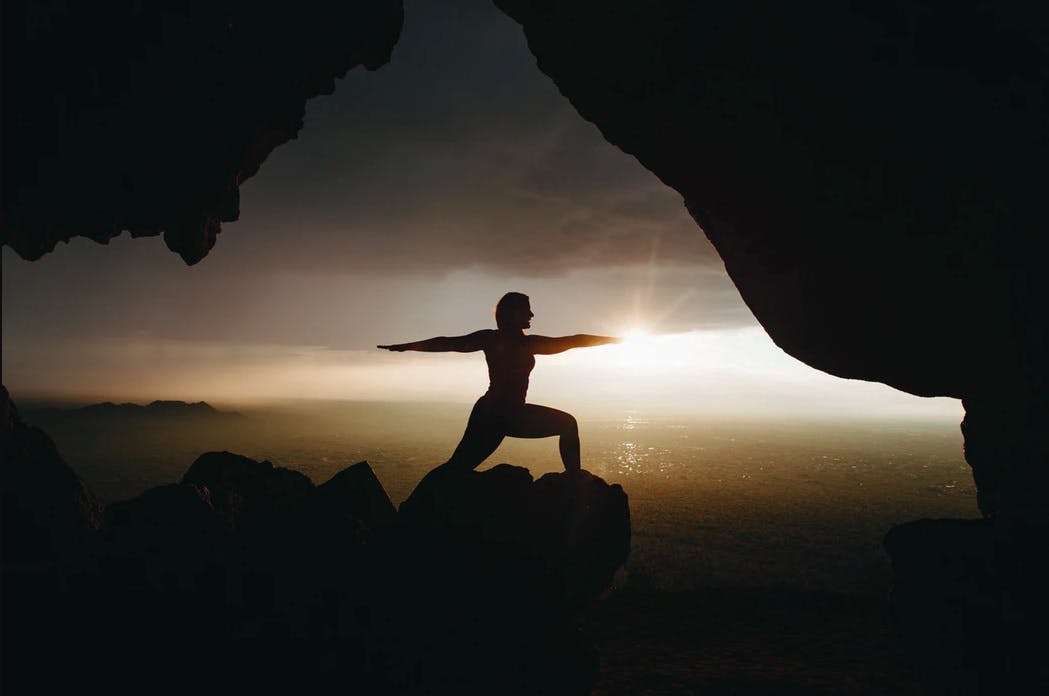 Silhouette of person in yoga posture on top of cliff during sunset