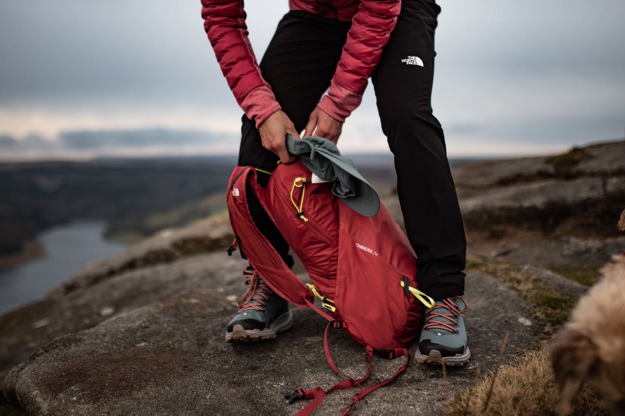 Marie öffnet ihren The North Face Chimera 24 L Rucksack auf einem Felsvorsprung im Hintergrund der Peak District.
