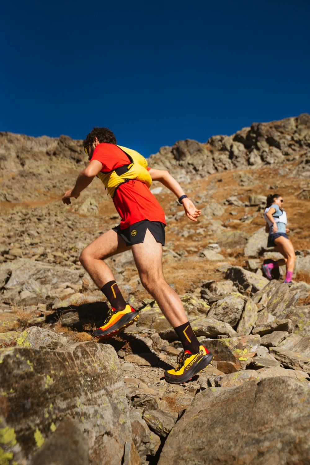 A trail runner in a red hydration vest sprinting up a steep rocky mountain path wearing La Sportiva performance accessories.